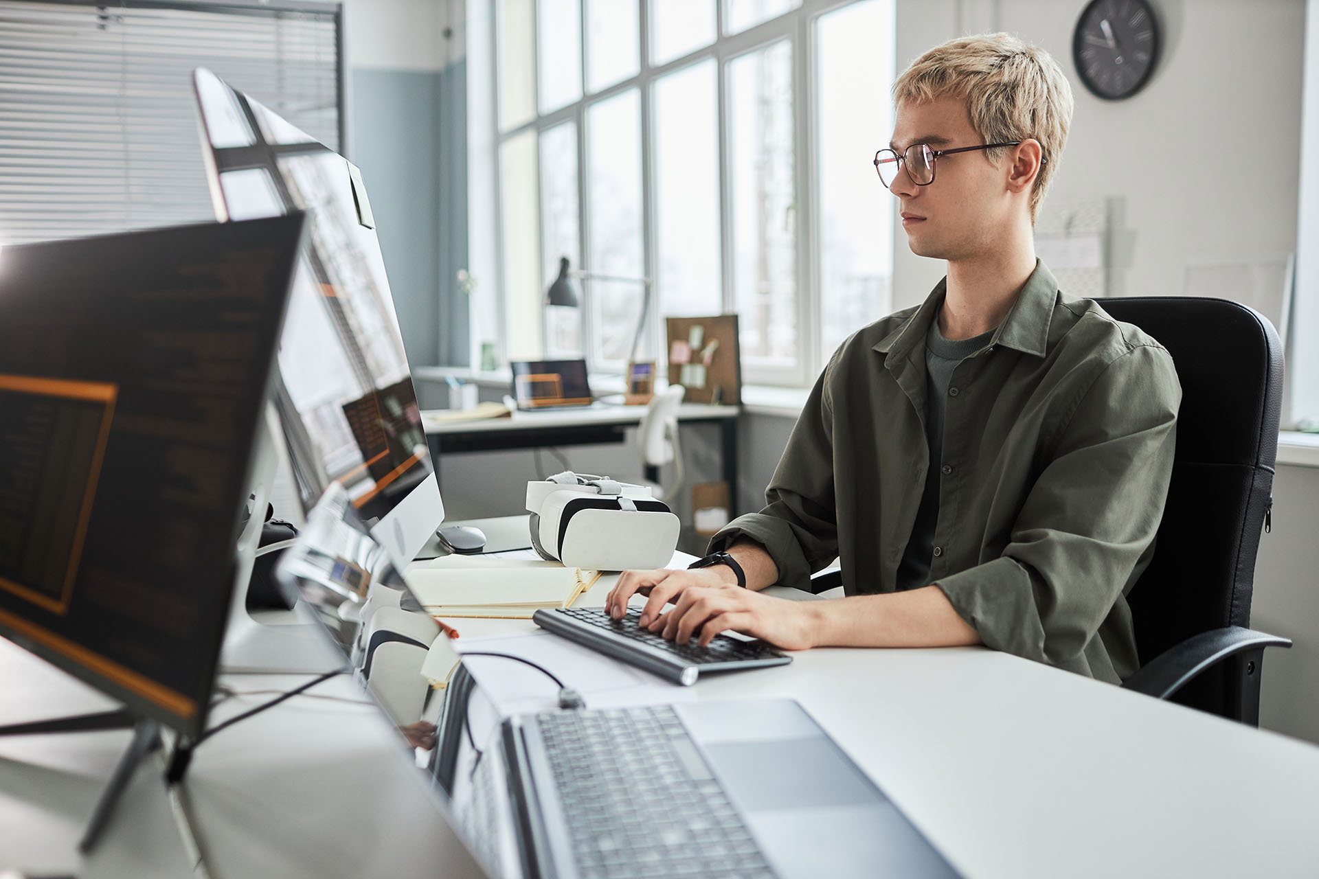 Young developer in eyeglasses writing codes for new software while sitting at his workplace in front of computer monitor